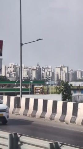 A vertical video shot from a moving vehicle on a highway. In the foreground, a concrete barrier with black and white stripes is visible. Beyond the barrier, a road with cars can be seen. In the middle ground, there is a green overhead structure that appears to be part of a highway or bridge, with another road visible underneath. A billboard advertising "TDI 10 Ludhiana" with an image of two men is mounted on a metal frame. Further in the background, a dense cluster of multi-story apartment buildings dominates the skyline under a hazy sky. A street lamp is also visible. The video pans slightly to the right.