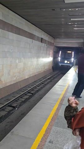 The video captures a subway train arriving at a station platform. The train, colored blue and white with horizontal red, blue, and green stripes, pulls into the station. The train's destination display reads "CHILONZOR" and the number "003" is displayed on the front. Passengers can be seen inside the train through the windows. A yellow safety line is visible on the platform edge. A person holding something in their hand stands near the camera. The subway station walls are composed of light brown stone tiles.