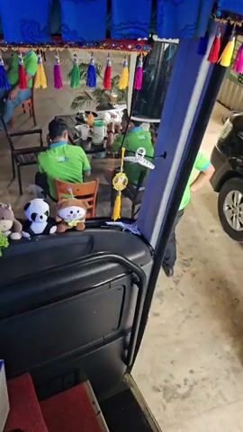 A video shows the interior of a bus, decorated with various stuffed animals and colorful tassels hanging from the ceiling. In the foreground, on the dashboard, are several stuffed animals including a blue bear, a panda, and a brown bear. Beyond the dashboard, two people in green shirts are seated at a table, appearing to be eating or talking. The background shows a seating area with chairs and tables, and the exterior environment is dimly lit, suggesting it is nighttime. The bus appears to be parked or stopped.