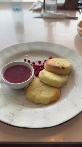 The video displays a close-up shot of a white plate holding a serving of syrniki, a type of fried quark pancake. Three syrniki are stacked on the right side of the plate, lightly browned with some darker spots from cooking. To the left of the syrniki, a small white ramekin is filled with a vibrant red berry sauce. Scattered between the syrniki and the sauce are a few small red lingonberries, used as garnish. The plate itself has a textured, speckled pattern. The syrniki are dusted with a light coating of powdered sugar. In the background, the setting appears to be a restaurant or cafe, with hints of a wooden table, a blurred glass of water, and a white napkin. The lighting is natural and soft, suggesting an indoor environment during the day. The camera angle is slightly overhead, focusing on the food.