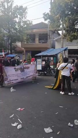 This video captures a street scene with a parade or procession. In the foreground, a large banner is held by several individuals. The banner is white with text and logos in various colors, including red, black, and gold. The text appears to be in Indonesian. Several people are visible behind the banner, some wearing traditional clothing, including headscarves and patterned skirts. Others are in more casual attire. To the right of the banner, a person in a traditional, ornate costume with a tall headdress is walking. Behind them, a motorcycle is parked. The street itself is asphalt and littered with white and colorful pieces of trash. In the background, there are buildings and trees. The overall atmosphere suggests a cultural event or celebration taking place outdoors during the daytime.