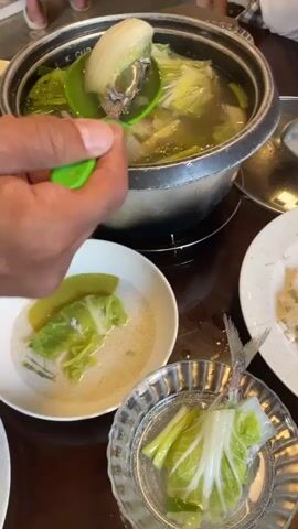 The video shows a table spread with a meal. At the center of the table is a cooking pot filled with soup, cabbage, carrots, and leeks. Someone uses a green spoon to serve the soup into a white bowl. To the right of the pot, a small clear plate holds a piece of fish wrapped in cabbage leaves. A larger white plate contains a mound of cooked rice with a serving spoon. A plastic container filled with stir-fried noodles and vegetables sits near the edge of the table. A coaster made of square wooden tiles is visible under the rice plate. The tabletop appears to be made of dark wood.
