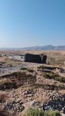 The video shows a wide view of a rural, hilly landscape under a clear blue sky. In the foreground, there is dry, yellowed grass and some scattered rocks. To the left and slightly further back, there is a ruined stone structure that appears to be a wall or part of a building, with some green vegetation growing on top of it. Next to this ruin, a small, white building with a green roof is visible. The middle ground consists of rolling, dry hills with some sparse vegetation and faint dirt paths winding through them. In the distance, more hills and mountains are visible, with a hazy appearance suggesting a long distance. The lighting indicates it is daytime, likely morning or afternoon. There are no people or animals clearly visible in the frame. The environment appears arid and somewhat undeveloped.
