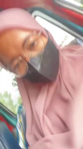 The video shows a close-up, selfie-style view of a person wearing a pink hijab, glasses, and a black face mask. The person is inside a vehicle, possibly a tuk-tuk, with a red and green interior visible. The camera angle is tilted, and the person's head is angled as well. In the background, through a window, foliage and a cloudy sky are visible. A hand briefly enters the frame and gestures near the person's face. The person's eyes are visible through their glasses, and they appear to be looking at the camera. The video is shaky, indicating movement of the vehicle.