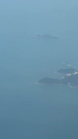 An aerial shot of a shoreline with a city and buildings on it, surrounded by water and islands, can be seen. The camera moves to the right.