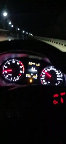 The video shows a car driving at night. The camera is positioned from inside the car, behind the steering wheel, showing the dashboard and the road ahead. The dashboard instruments, including the speedometer and tachometer, are visible and illuminated. The speedometer indicates the car's speed fluctuating between 69 and 78 km/h. The road is wet and illuminated by streetlights. There is a barrier along the right side of the road. No people are visible, but the presence of a driver is implied by the perspective. The overall environment appears to be a highway or major road.