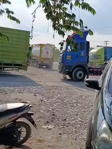 The video shows a blue semi-truck with a white tanker trailer attached, moving slowly on a gravel road. The truck is on the right side of the frame, and the trailer extends to the left. In the foreground, on the left, a portion of a motorcycle is visible. To the right of the motorcycle, a black car is partially visible, obscuring the right side of the truck and trailer. In the background, other trucks are parked or driving, and trees and foliage are present. The sky is cloudy. The video appears to be taken from a stationary position, possibly from inside the black car. The truck is the primary focus of the video, and its movement is the main action.