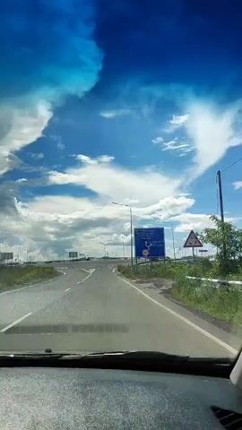 The video shows a view from inside a car driving on a highway. The sky is blue with scattered clouds. There are road signs indicating directions and speed limits. The road has multiple lanes and curves to the right. The scenery outside the car appears to be grassy fields. The car's dashboard is visible in the foreground.