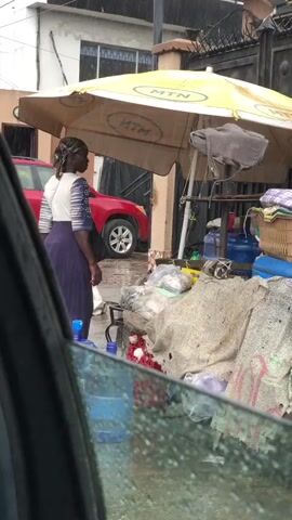 A woman wearing a purple skirt and a patterned white and blue shirt stands under a yellow umbrella that displays the MTN logo. She is facing right, holding a bag and interacting with a vendor stall. In front of her, a collection of items is laid out on a tarp, including blue water jugs, some plastic bags with contents, and what appears to be food items. Behind her, a red SUV is parked. The ground appears wet, suggesting it may be raining, and the overall environment seems to be an outdoor market or roadside stall.