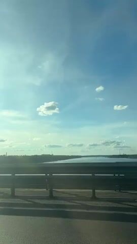 The video shows a view from a moving vehicle on a bridge. The sky is blue with scattered white clouds and the sun is shining brightly. In the distance, a body of water is visible, with trees and buildings on the far bank. A metal guardrail runs along the side of the bridge, casting shadows on the asphalt road. A person with a backpack is seen standing on the bridge, leaning on the guardrail and looking out at the view. The camera is angled slightly upwards, capturing the sky and the distant landscape.
