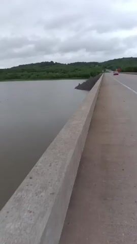 The video opens with a view from a concrete bridge, showing a railing to the left and a road to the right with a car visible in the distance. The camera then pans left to reveal a wide, murky brown body of water under an overcast, cloudy sky. Distant forested hills line the shores of the water on both sides. Small specks, possibly debris or birds, are scattered across the water's surface, and one larger white object is briefly visible floating in the water. The scene depicts a serene, albeit cloudy, landscape, likely a lake or wide river, in Cookson, United States.