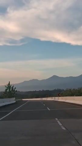 The video shows a highway with mountains in the background and a clear blue sky with some clouds. The road is paved and has white lane markings. There are also some trees and bushes on the side of the road. The video appears to have been filmed from a car traveling on the highway. The lighting suggests it is either early morning or late afternoon.