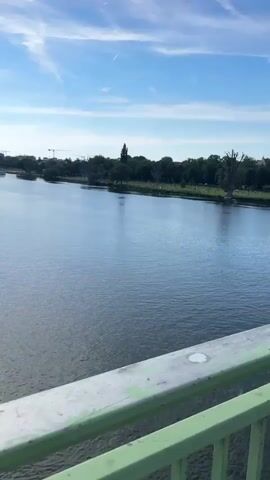 The camera, positioned on a bridge, overlooks a wide river with a green park on the opposite bank, under a blue, partly cloudy sky.  The river’s surface is relatively still, reflecting the sky. On the far bank, the green park is dotted with numerous indistinct figures, suggesting people relaxing or engaging in activities. A thin, white contrail stretches across the sky. The bridge’s railing, painted a weathered green, runs across the foreground. The footage is steady, capturing the serene scene of urban nature.