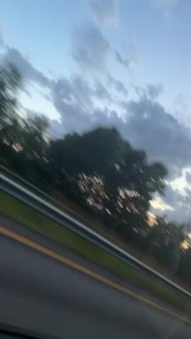 This is a blurry video of trees and clouds passing by from the perspective of a moving vehicle. The sun appears to be setting, casting a warm glow on the clouds and trees. A yellow line on a road is visible in the foreground.