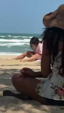 Two women are sitting on a sandy beach. The woman closest to the camera is sitting cross-legged, wearing a straw hat with a bow and a floral-patterned top. She appears to be holding something thin in her hand, possibly a cigarette. Behind her, another woman is lying on the beach, looking at a phone. The ocean with small waves is visible in the background under a clear sky.