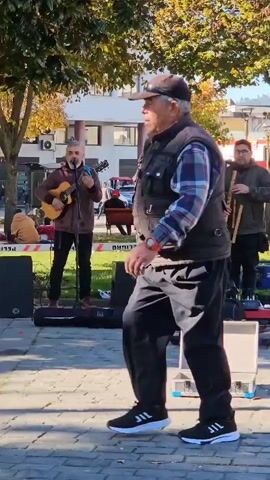 A group of four men are performing as a band in a park. One man is playing a guitar and singing into a microphone, another is playing a charango, and a third is playing a wooden wind instrument. The fourth member has joined the band and is setting up his saxophone. They have a white case on the ground which may be used for receiving tips, along with other music equipment. People are walking by, listening to their performance. A woman and her friend stop to record the performance on a cell phone. The weather appears to be sunny and the park is surrounded by buildings. The scene shows a typical day in the park with music filling the air.