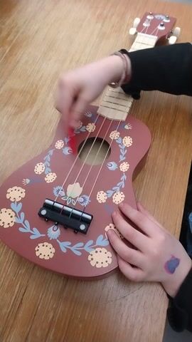 A child sitting at a wooden table strums a brown toy ukulele, decorated with a floral design. The ukulele has a light-colored fretboard and black bridge. The child wears a black long-sleeved shirt and a blue shirt underneath. A red stick is used to strum the strings. The child has a reddish-purple mark on the back of their left hand. The table is made of light wood.