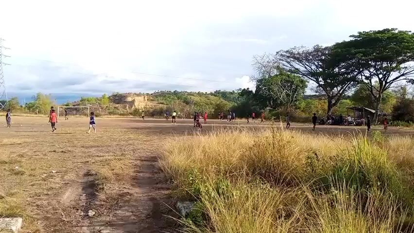 A group of people, likely children, are playing soccer on a dry, grassy field under a cloudy sky. The field is uneven and dusty, with a few scattered trees in the background. Some spectators are gathered under a makeshift shelter on the right side of the frame. The scene appears to be in a rural or undeveloped area. The camera is positioned at a low angle, giving a sense of looking up at the players and the environment.