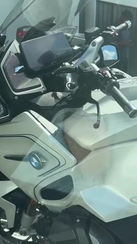 This is a close-up, eye-level, medium shot of a white BMW motorcycle parked outdoors. The motorcycle has a silver and blue emblem with the BMW logo on its fairing, and the model designation "R1250RS" is visible. The motorcycle's dashboard and handlebars are also visible, as is part of the front wheel with disc brakes. The sun is shining brightly, casting shadows on the motorcycle and the surrounding environment, which appears to be a street or parking lot with a building in the background. There are a few people visible in the background, but they are out of focus. The video appears to be shot from a vehicle moving slowly or stopped.
