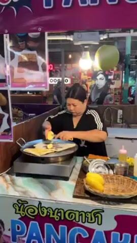 A woman is making a pancake at an outdoor food stand. She is using a yellow knife to cut up a mango on top of the pancake. The pancake is on a round hot plate. The woman is wearing a black shirt with white stripes down the sleeves. The food stand is decorated with posters and lights. There is a wicker basket with mango in it on the counter next to the hot plate.