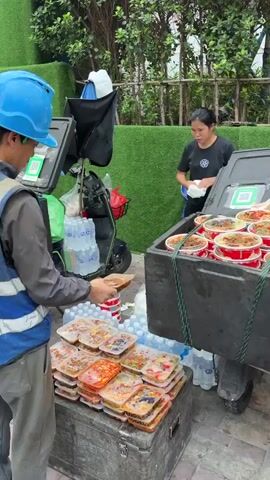 This media file captures a busy street scene in the Nanshan District of China, likely during the daytime given the lighting. The primary focus is on the exchange of food items between several individuals.

In the foreground, a person wearing a blue hard hat and a reflective vest is attending to stacks of food containers. Nearby, a woman in a black t-shirt is holding a white paper bag and appears to be involved in the food distribution. A large, dark cooler, filled with numerous round food containers, is prominently featured. Another individual, wearing a yellow hard hat and a bright yellow reflective vest, stands to the right, seemingly observing or waiting. In the background, there's a scooter loaded with water bottles and bags, suggesting logistics or delivery. The setting includes a green fence or wall, foliage, and a street or pathway.

The activity depicted is the organized distribution or sale of pre-packaged meals. The sheer volume of identical containers implies a catering or food delivery service, potentially for workers at a nearby construction site, as indicated by the hard hats and reflective vests. The presence of multiple people engaged in the process suggests a coordinated effort.

Notable details include the abundance of food containers, both in the cooler and stacked on a wooden crate. QR codes are visible on the cooler, likely for payment or identification. The people appear focused on their tasks, with no overtly strong emotions displayed, suggesting a routine operation. The time of day is likely daytime, as natural light illuminates the scene. The presence of construction scaffolding in the far background provides a clue about the broader environment.

Visible text includes Chinese characters on the yellow vests of the individuals, and potentially on signage in the background which is partially obscured. The QR codes are also a form of visual information.