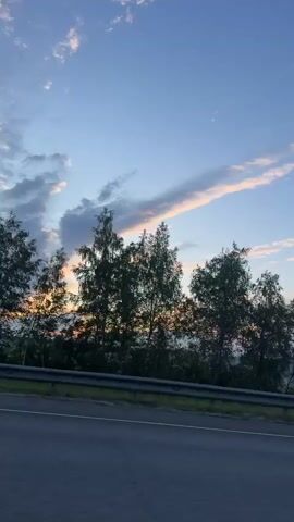 A video captures a view of a road and trees against a blue sky with clouds. The camera appears to be mounted on a moving vehicle, suggesting the shot is taken from a car or bus. The sun is setting, casting a warm glow on the clouds. The trees are silhouetted against the sky, and their branches are visible. The road is asphalt, and there is a guardrail on the side. The overall impression is one of a serene and peaceful landscape.