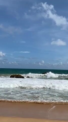 A video shows a beach with waves crashing on the shore. The sky is blue with white clouds, and the ocean is a deep green. There are a few rocks in the ocean, and the waves are breaking over them. The sand on the beach is a light brown color. The video is shot from a low angle, so the viewer is looking up at the sky and the ocean.