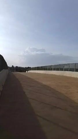 The camera is static, facing down a wide, empty, light-brown colored walkway that runs parallel to a long, low fence constructed of vertical metal bars mounted on a white concrete base. A single figure walks away from the camera down the walkway. The sky is blue with a large white cloud hovering above the horizon. The sun appears to be behind the camera, as long shadows stretch across the walkway towards the horizon.