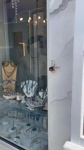 The video shows a storefront display featuring jewelry and scarves. The display is divided into multiple shelves. The top shelf has sunglasses and necklaces. The second shelf has necklaces and bracelets. The bottom shelf has a variety of jewelry, including necklaces, bracelets, and earrings. To the right of the jewelry display are folded scarves with patterns of fish and other designs. Further to the right, there are racks of clothing and hats. In the background, through the window, a manikin is visible wearing a necklace. The scene then shifts to a scenic view of a coastal town with white buildings and the blue sea. The water appears calm, and there are boats visible in the distance. The sky is clear and blue.