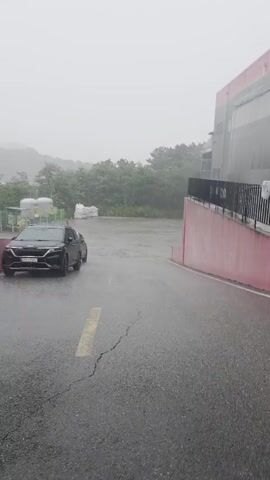 A black SUV is parked on a wet, asphalt road, under a light gray sky. Heavy rain is falling, and the road is covered in puddles. To the left, a red barrier runs along the side of the road. Behind the car, there are several white storage tanks and some greenery. To the right, the edge of a building with a black railing is visible. The scene appears to be an industrial or commercial area.