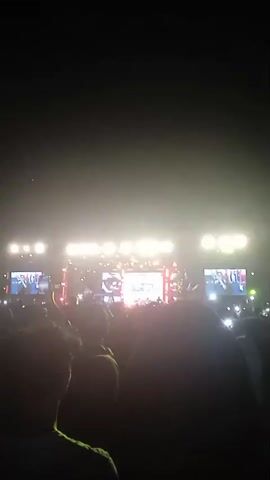 This is a vertical video of a night-time outdoor concert. The scene is dominated by bright yellow and white stage lights that illuminate a large crowd of people gathered in front of a stage. The stage has multiple large screens displaying what appear to be performances or visuals, with a central screen and two flanking screens. The crowd is a dark silhouette against the bright lights, with many people holding up their phones to record or take pictures. The overall mood seems to be energetic and festive, typical of a concert. The time of day is clearly night due to the dark sky and the reliance on artificial lighting. There are no clear weather cues, but the atmosphere suggests a clear night. The location context provided is Kendal, Indonesia, suggesting this is a public event in that region. No discernible text is visible in the video.