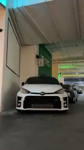 The video shows a white Toyota GR Yaris parked in an indoor parking garage. The car is facing the camera and is slightly angled to the left. There is another car parked behind it. A sign for "The Pulse Kitchen" is visible on the wall next to the car. The lighting in the garage is cool and artificial, with fluorescent lights on the ceiling. The floor is concrete and appears to be clean. There are no people visible in the video.
