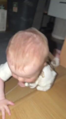A baby is on its stomach on a wooden floor, reaching forward with its left hand. The baby is wearing a light-colored onesie with a pattern of bears. The background is dark and out of focus, showing what appears to be storage boxes and furniture. The baby's head is turned to the left, and its eyes are open.