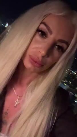 This media file is a close-up, first-person perspective video of a woman with long, blonde hair, wearing a dark jacket and a cross pendant necklace. Her makeup is noticeable, with bold eyeliner and full lips. The background appears to be an outdoor setting at night, with city lights visible in the distance. There are out-of-focus figures and lights in the background, suggesting a public or social gathering. The lighting is dim, characteristic of evening or nighttime. The woman is looking directly at the camera and her expression is neutral, possibly with a slight pout. No visible text is present. The context provided indicates the location is Khan Boeng Keng Kang, Cambodia, suggesting an urban nighttime environment.