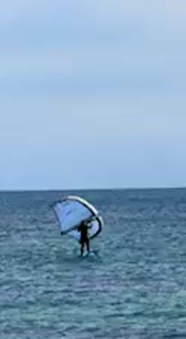 A person is captured in a long shot, standing on a board in the open sea, engaged in wing foiling. They are wearing dark attire and holding a light-colored wing (a sail-like apparatus) above their head. The wing is predominantly white or light blue, with some darker accents. The water is a deep blue-green, showing slight ripples. The horizon line is visible, separating the ocean from a pale blue-grey, overcast sky. The overall scene suggests a calm day, possibly with moderate wind, suitable for water sports. The setting is the sea off Barbate, Spain.