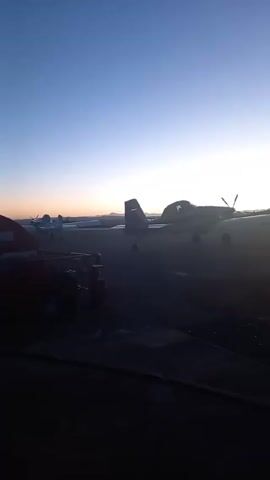 The video shows several planes parked on a runway, likely at dawn or dusk. The sky transitions from deep blue to lighter orange and pink as the sun rises or sets. The planes are silhouetted, making it difficult to discern details. A blurry object, possibly a vehicle or structure, is visible in the foreground on the left. The ground appears to be a combination of paved areas and darker, possibly gravel, surfaces. Overall, the scene evokes a sense of quiet and anticipation, with the planes ready for flight at the beginning or end of the day.