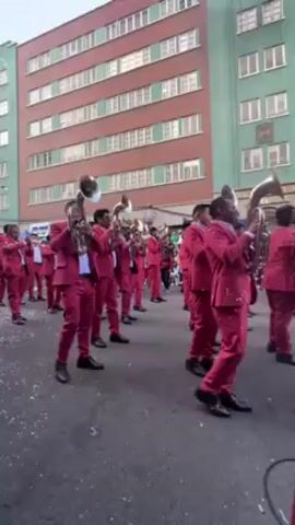 A street parade features a brass band in matching pink suits. The band members are lined up playing their instruments. Passersby walk in front of them, including a woman in traditional Andean clothing and other pedestrians. Confetti covers the ground. Tall buildings line the street.