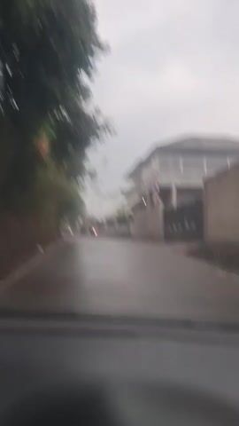 The video, shot from inside a vehicle, shows a wet road with trees and vegetation on the left side and buildings on the right. Two motorcycles are seen traveling on the road. The weather appears overcast and possibly rainy. The video progresses to show more of the road and the surrounding environment as the vehicle moves forward.