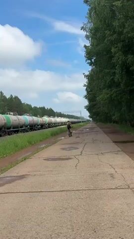 A person rides a bicycle down a paved path beside a long train of tank cars. The train cars are white and green and extend far into the distance. The path is cracked and has puddles of water on it. To the right of the path is a dense line of trees. The sky is blue with scattered white clouds.