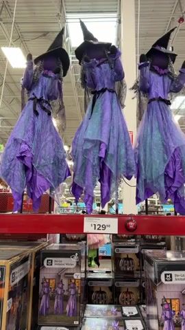 The video shows three hanging witch decorations for sale in a retail store. The witches are dressed in purple and green iridescent gowns and black pointed hats. They are displayed on a shelf above other merchandise, including boxes with images of Halloween decorations. A price tag of $129.98 is visible below the witches. The background appears to be a typical retail environment with high ceilings and fluorescent lighting.