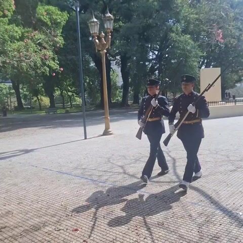 Two individuals in dark blue uniforms and caps march in a park. They are carrying rifles and wear yellow belts with leather pouches. The park has trees, benches, and a paved ground with a checkered pattern. A lamppost is visible. The scene is bright and sunny, with shadows cast by the trees and the individuals.