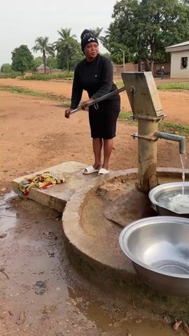 A woman operates a manual water pump in a rural outdoor setting in Tabu, Nigeria. She stands on a concrete platform, wearing a black beanie with a white logo, a black long-sleeved top, a black skirt, and white sandals. Her expression is neutral and focused as she works the pump handle.

Water streams from the pump's spout into a large, silver metal bowl already partially filled with water. A second, empty silver metal bowl sits next to it, presumably waiting to be filled. Colorful fabric or clothes are folded on the concrete platform beside the woman.

The ground around the pump is dry, reddish-brown dirt, with some muddy patches directly beneath the spout. In the background, there are several green trees, including tall palm trees, and a simple, light-colored building. The lighting suggests it is daytime, with soft, diffused light.