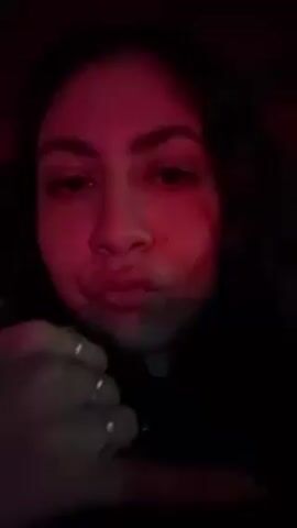 A young woman appears to be filming a video. She is indoors, presumably at night, under red lighting. She is looking directly into the camera with pursed lips. She is wearing rings on her fingers and a black top. She makes a hand gesture. The background is dark, and music is audible.