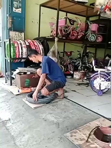 A man is squatting on the ground, working on a tire. He is wearing a blue t-shirt and dark shorts. In front of him is a tire laying on a piece of cardboard. He appears to be using his hands to work on the tire. Behind him, there are shelves stocked with tires and various bicycles. The scene appears to be an outdoor workshop or repair shop. The ground is concrete and there is some debris scattered around. The lighting suggests it is daytime.