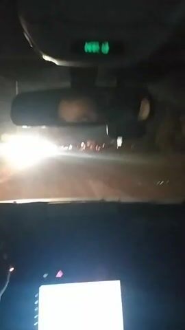 The video shows a view from inside a car at night. The dashboard and rearview mirror are visible. Through the windshield, one can see headlights and taillights of other vehicles on a road, as well as some lights from nearby buildings. The dashboard display indicates it is March 13th. A person's face is visible in the rearview mirror. The car appears to be in motion.