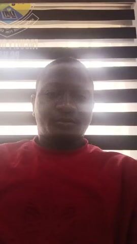The video shows two dark-skinned men in an indoor setting. The first man is wearing a red shirt and is the primary focus in the initial shot. He is looking directly at the camera. The second man is wearing a striped shirt with red, white, and blue bands, along with a gray section featuring a large "LV" logo. He is visible in subsequent shots, looking to his side. The background includes a wall and a partially visible window with horizontal blinds that are letting light in. The camera angle changes, shifting from a straight-on view to a low-angle shot looking upwards at the second man. There is no significant action or interaction captured in the video.