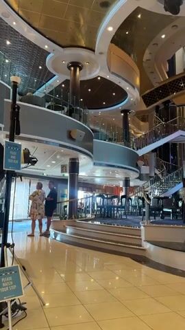 This video captures the interior of a grand atrium, likely on a cruise ship or in a large hotel. The scene is characterized by its opulent design, featuring multiple levels, ornate railings, sweeping staircases, and a dramatic, multi-tiered ceiling with golden accents and recessed lighting. A striking, abstract metallic sculpture is centrally positioned in the foreground.

The atrium appears to be a communal space, possibly a lounge or dining area. Several tables and chairs are visible on different levels, suggesting it's designed for people to gather and dine. Two individuals are standing near the bottom left of the frame, seemingly engaged in conversation. The lighting suggests it is daytime, or at least a well-lit interior, with natural light possibly filtering in from unseen windows or balconies on upper levels. The polished floor reflects the bright lights and architectural features, adding to the sense of spaciousness and luxury.

There's a sign visible on a stand in the lower left corner of the frame with the text "PLEASE KEEP YOUR HANDS OVER YOUR HEAD". The overall atmosphere is one of sophistication and grandeur, hinting at a high-end establishment. The location cue "Ferragudo, Portugal" suggests this grand interior is part of a resort or facility in that Portuguese town. The scene is static, focusing on the architectural marvel and the ambiance it creates.