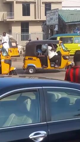 The video captures a daytime street scene in Dadi Riba, Nigeria. A large herd of white cows, some with darker markings, is being herded across a busy road, causing vehicles and pedestrians to navigate around them.

In the foreground, a black car is partially visible on the left, with a man in a red and black striped shirt standing beside it, facing away from the camera. In the middle of the road, a yellow auto-rickshaw (keke) carrying passengers moves slowly past the herd. A white motorcycle also rides through the street, and a green and yellow bus is briefly visible. On the left side of the road, a man in a white robe stands near a fence, observing the scene, with one of the cows close by.

Buildings line both sides of the street, including multi-story structures and ground-level shops. One shop displays various items and features a sign that partially reads "CENERAL PURSHING CO." The weather appears clear and sunny.
