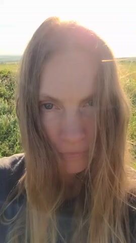 A woman with long blonde hair and blue eyes is shown in a close-up video. The video begins with her looking directly at the camera with a neutral expression. She then blinks slowly and squints her eyes. The background appears to be an outdoor setting with green grass and a bright sky, suggesting it is daytime. The sun is shining, creating a lens flare effect in the top of the frame. The woman is wearing a dark-colored shirt.