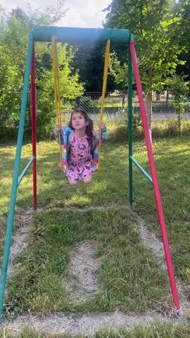 A young girl with dark hair is swinging on a playground swing. She is wearing a pink dress with a Minnie Mouse print and a colorful striped cardigan. She also has on pink sandals. The swing set is green and red with yellow chains. The girl is smiling and appears to be enjoying herself. The background shows trees and grass, suggesting an outdoor setting. The time of day appears to be late afternoon, with the sun casting shadows. The weather seems pleasant. There is no visible text in the image. The location is Botfalva, Ukraine.